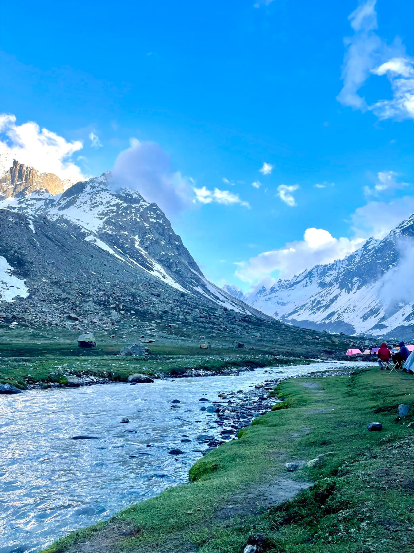 Tents beside a glacial river in a green valley, snow peaks behind
